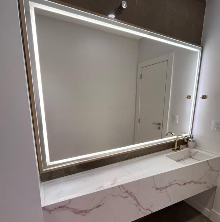 Espelho retangular com moldura dourada e iluminação LED em toda a borda, sobre bancada de mármore branco