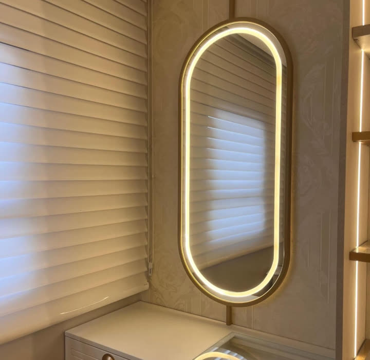 Espelho oval com moldura dourada e iluminação LED contornando a borda, instalado sobre um móvel claro