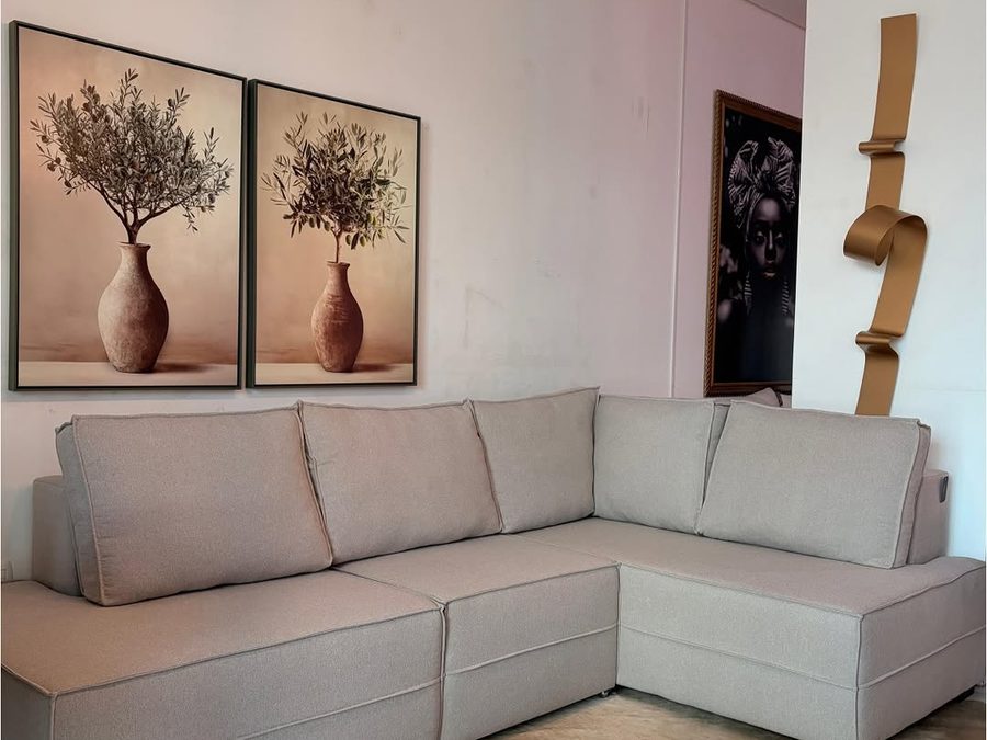 Sofá de canto em tecido bege montado no showroom, com quadros e luminárias pendentes ao fundo
