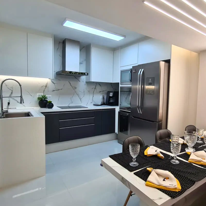 Cozinha em L com armários superiores brancos, bancada e gabinetes inferiores em grafite fosco, revestimento de mármore claro e iluminação linear no teto