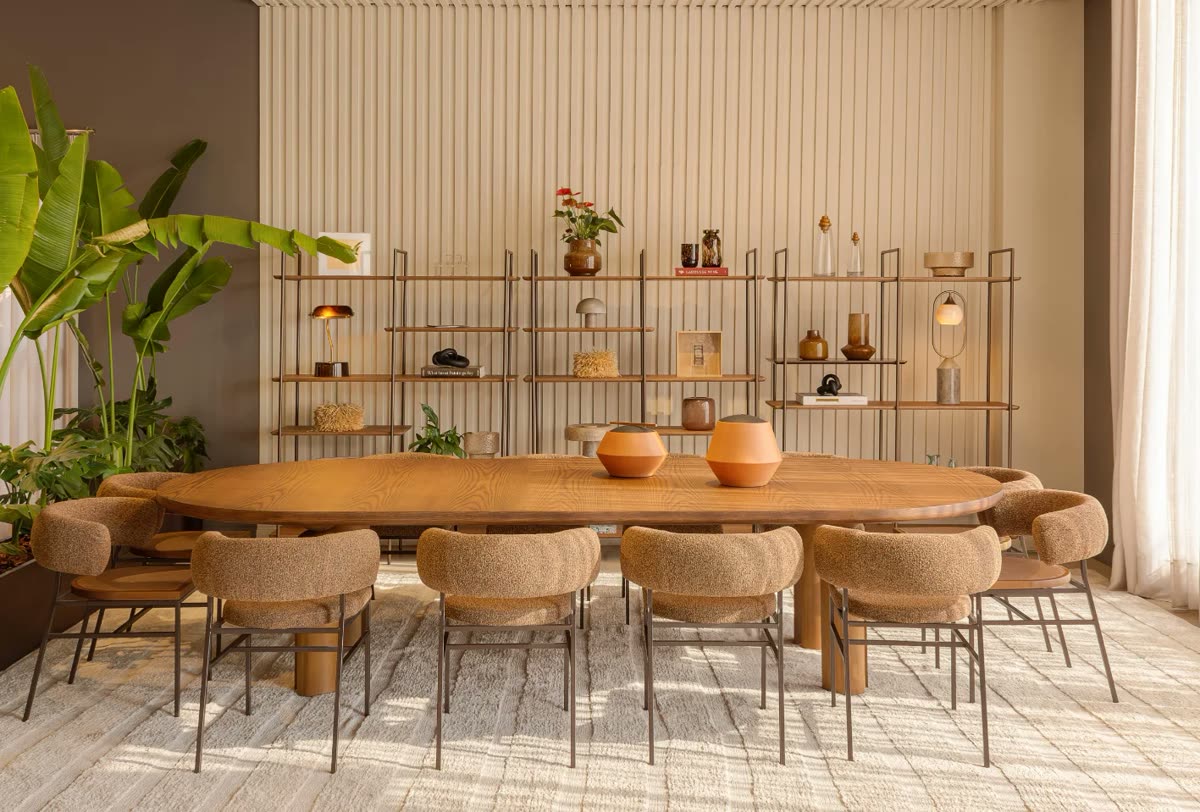 Mesa de jantar oval de madeira com dez cadeiras de bouclê caramelo, diante de estantes vazadas com objetos de decoração