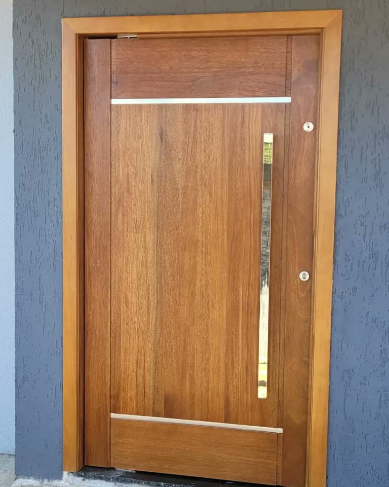 Porta de entrada de madeira maciça com puxador vertical em inox, instalada em fachada de textura cinza